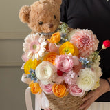 Load image into Gallery viewer, A person holds a decorative flower basket with a teddy bear head attached at the top. The bouquet includes pink, peach, yellow, white, and light blue flowers arranged in a woven basket with a white ribbon.